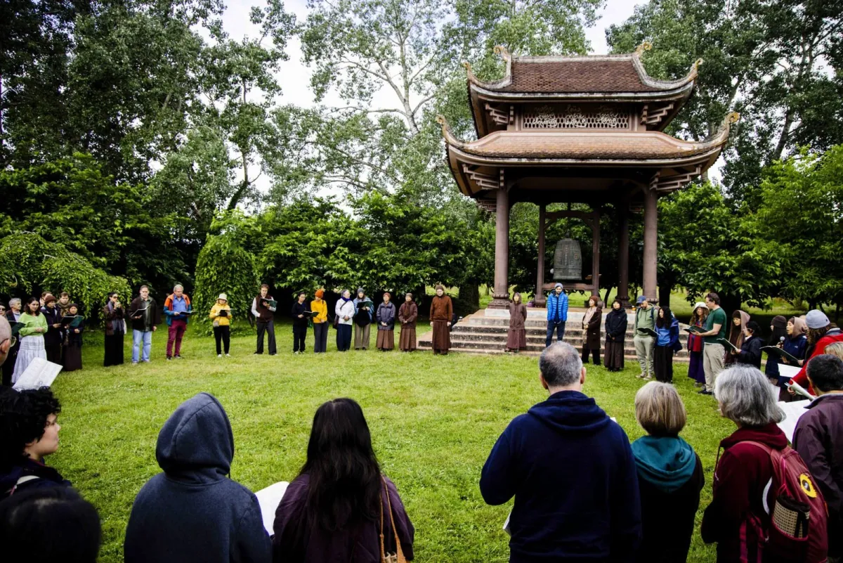 A wide circle of friends standing on the grass in front of the bell tower at Plum Village, songbooks in hand, trees all around.
